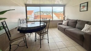 A living room with a dining table and chairs, a sofa, a large window with a view of buildings, and a framed picture on the wall. - A052 Cosy studio near Croisette and the beach (Cannes)