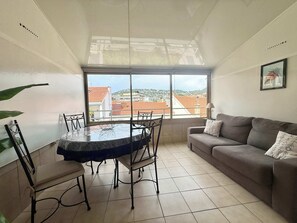 Ein Wohnzimmer mit Esstisch und Stühlen, einer Couch, einem großen Fenster mit Blick auf Gebäude und einem gerahmten Bild an der Wand.