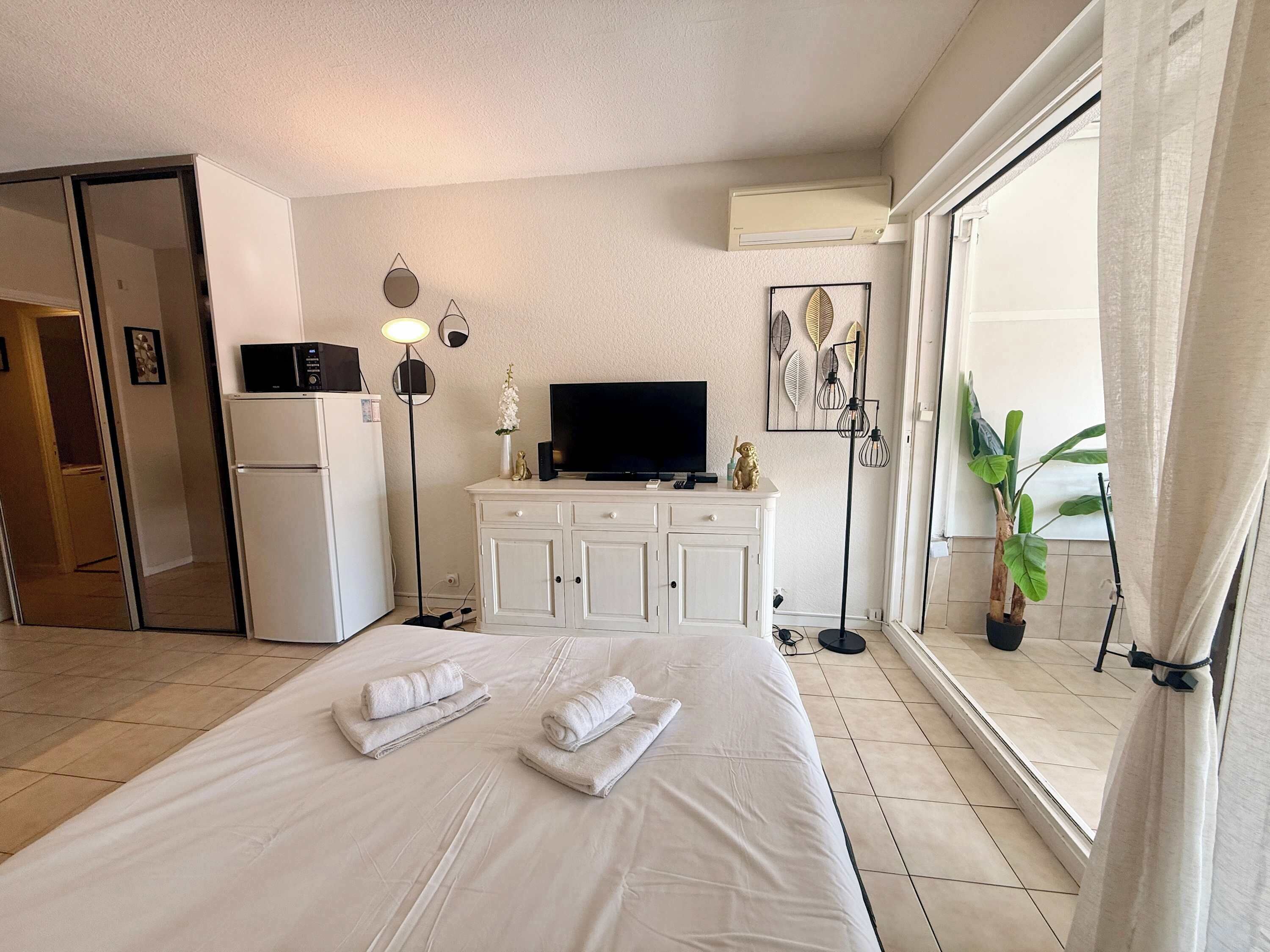 A hotel room with a bed, a television, a minibar, a desk, and a large window with curtains.