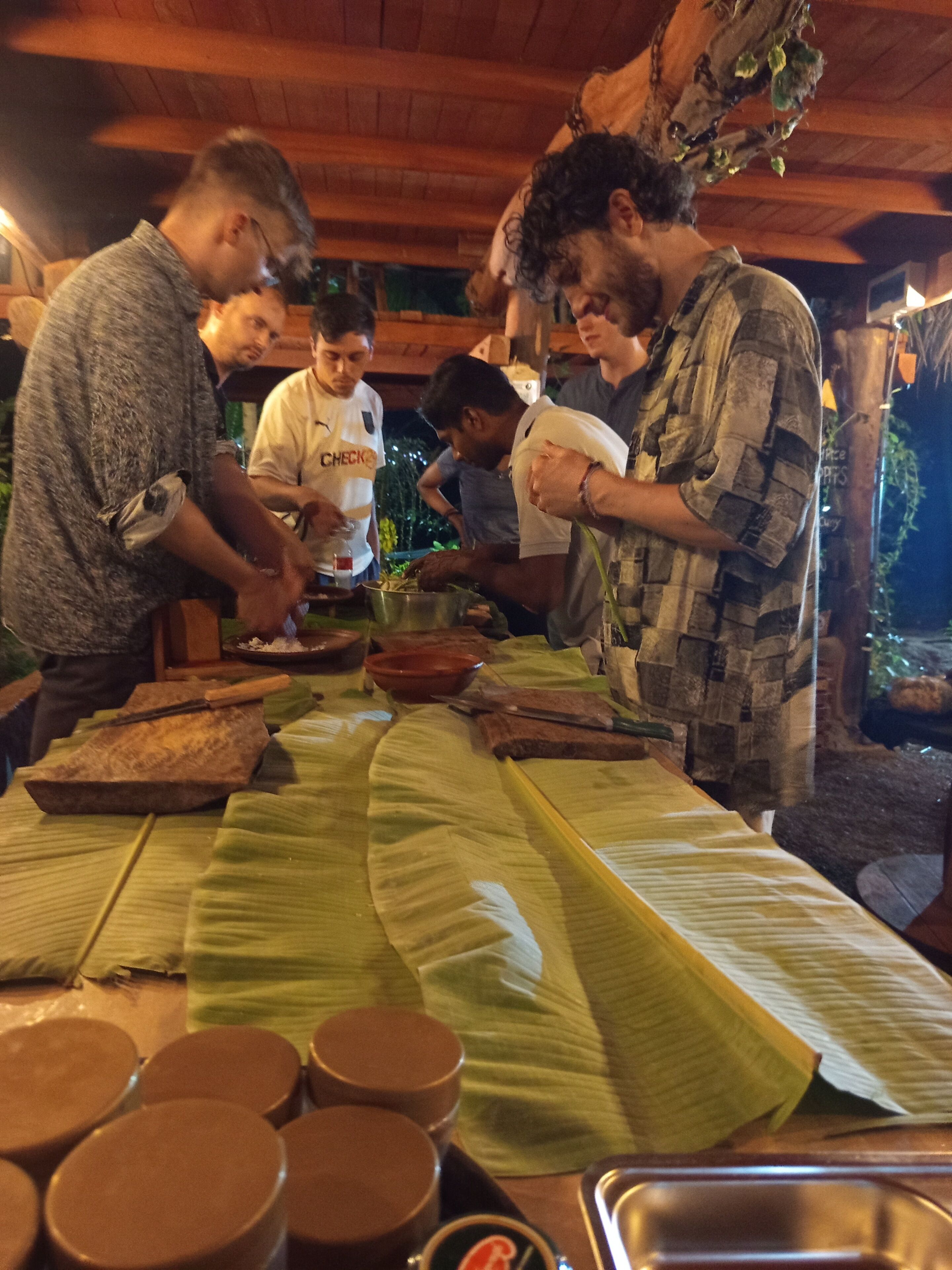 Een groep mensen bereidt eten voor aan een houten buitentafel met een groene tafelkleed, omgeven door houten balken en een rustieke omgeving.