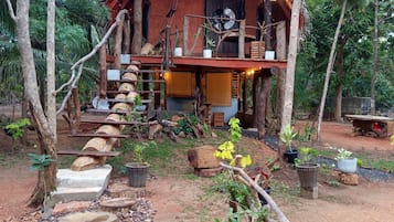 A rustic wooden house with a thatched roof, surrounded by trees and a stone pathway leading to the entrance.