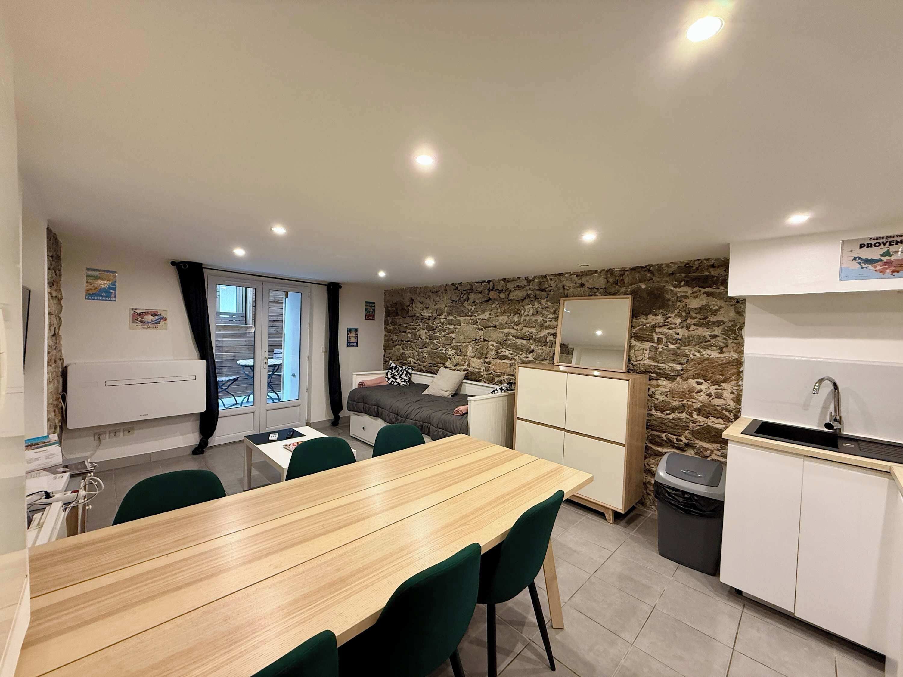Une pièce compacte comprenant une table à manger en bois, des chaises vertes, un lit avec un mur en pierre en arrière-plan, une petite kitchenette et une porte-fenêtre coulissante donnant sur un espace extérieur.