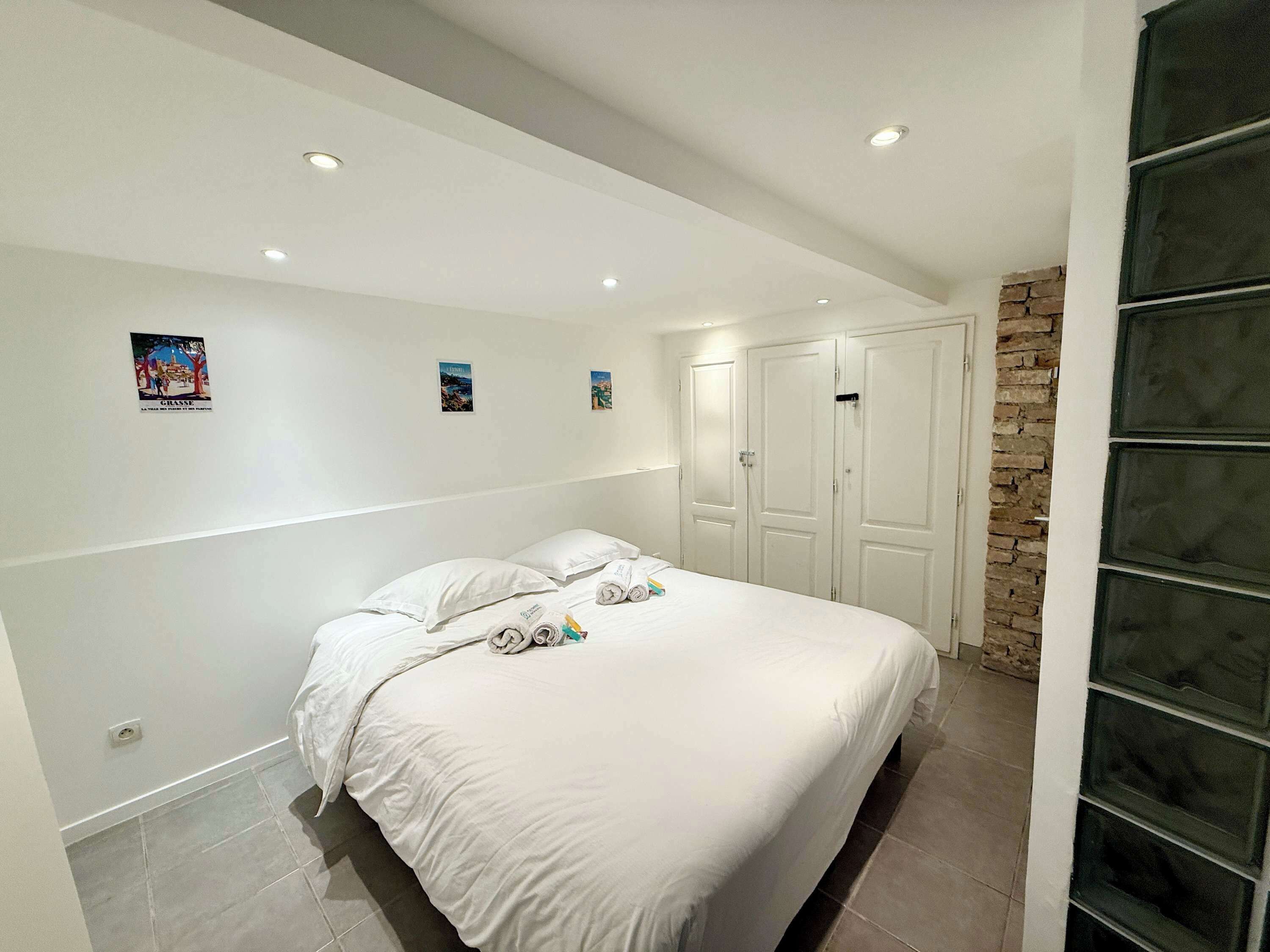 A bedroom with a large bed, white walls, and a brick accent wall. There are three doors and a small window.