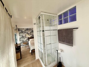 A room with a glass shower enclosure, a bed with a patterned headboard, a small table with a plant, and a wall with a blue-tinted window. - A088 Élégant duplex avec terrasse, plages, centre (Cannes)