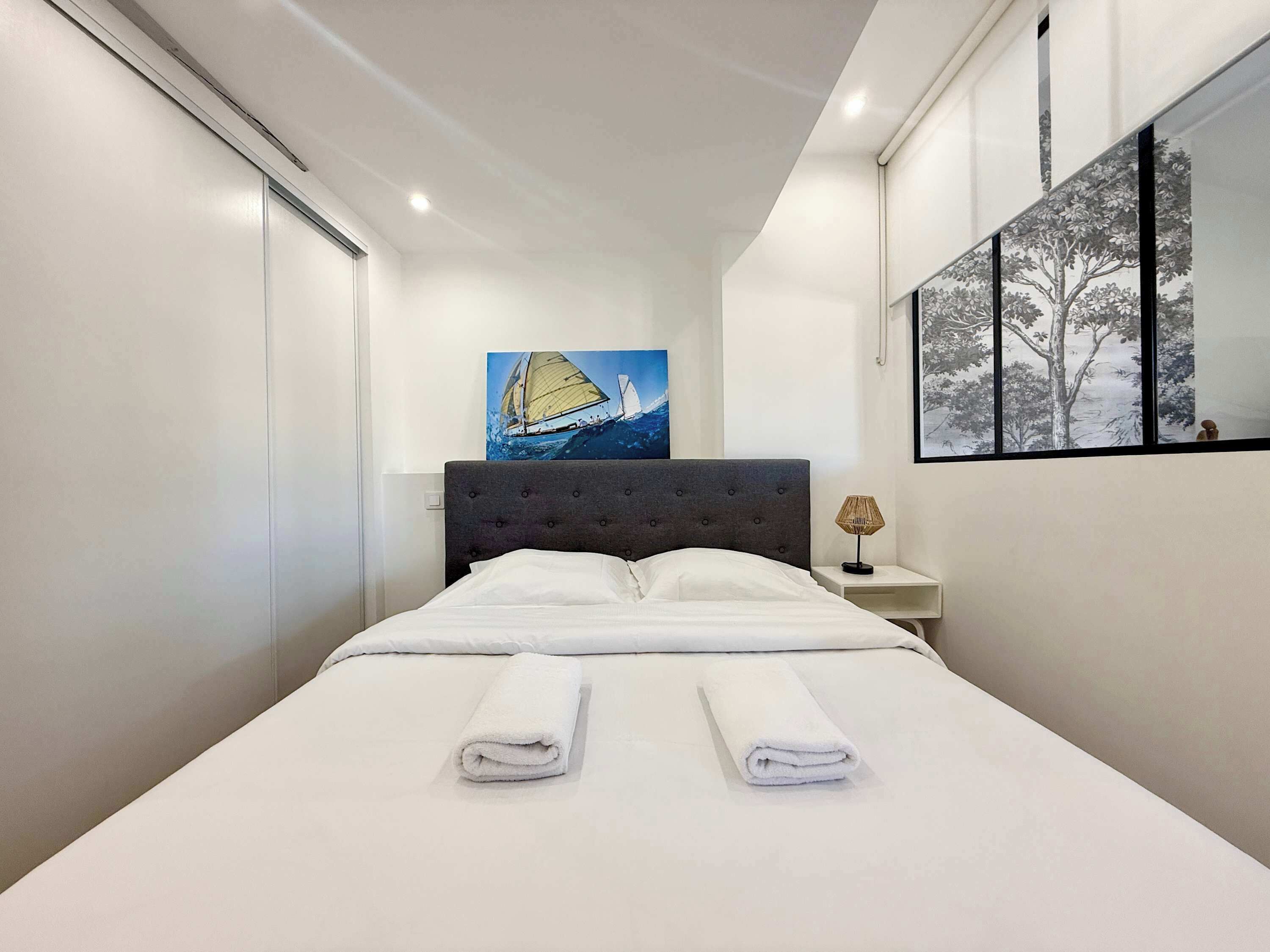 A modern hotel room with a large bed, a painting of a sailboat, a nightstand with a lamp, and a window with a view of trees.