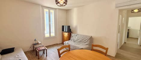 A room with a wooden dining table, a sofa with black cushions, a television on a wooden cabinet, and a window with curtains.