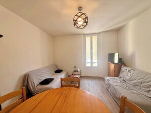 A small, well-lit room with a single bed, a sofa, a table, and a TV on a wooden cabinet. - C033 Appartement fonctionnel au coeur du Suquet (Cannes)