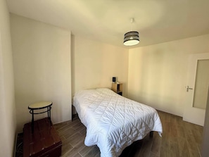 A bedroom with a bed, a small table, a chair, a lamp, and a door. - C033 Appartement fonctionnel au coeur du Suquet (Cannes)