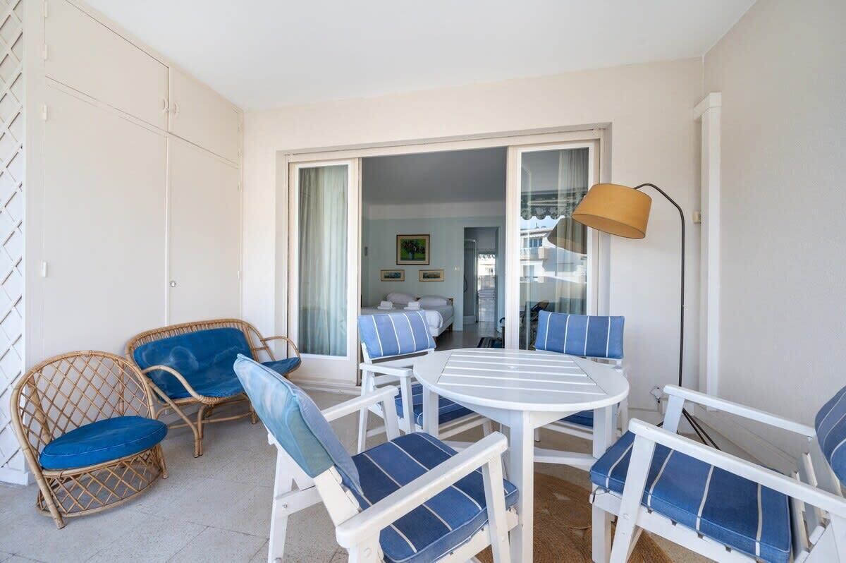 Un balcon avec des fauteuils en osier et une table blanche, une pièce avec un lit et un tableau, et une lampe sur la droite.