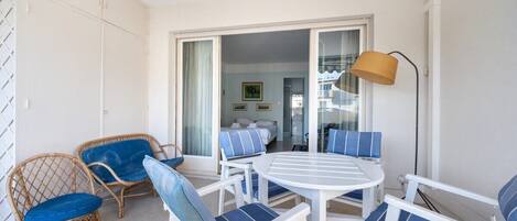 A balcony with wicker chairs and a white table, a room with a bed and a painting, and a lamp on the right.