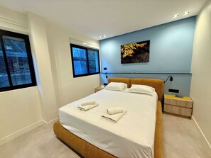 Ein modernes Schlafzimmer mit einem großen Bett, einem Nachttisch, einem Wandbild und einem Fenster mit Blick auf ein Gebäude.