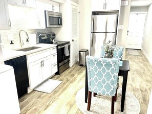 A modern kitchen with white cabinets, a stainless steel refrigerator, a microwave, and a dishwasher. There is a dining table with chairs and a small potted plant.
