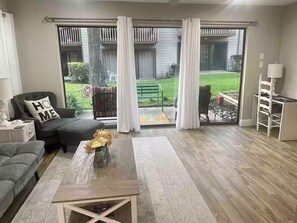Ein Wohnzimmer mit grauem Sofa, einem Couchtisch und Blick auf einen Garten durch eine Schiebetür.