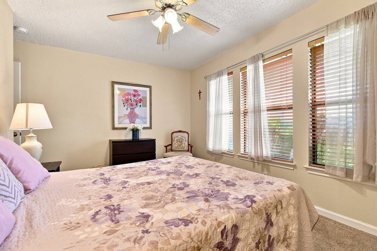 A bedroom with a bed, a nightstand, a lamp, a chair, a ceiling fan, and a window with blinds.