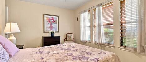 A bedroom with a bed, a nightstand, a lamp, a chair, a ceiling fan, and a window with blinds.