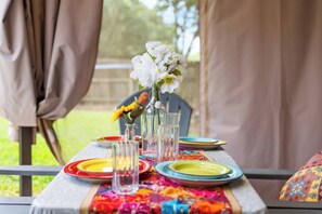 Ein Esstisch ist gedeckt, mit bunten Tellern, klarem Glas und einer Tischdecke mit Muster. Ein Blumenstrauß mit weißen Blüten steht in einer Vase.