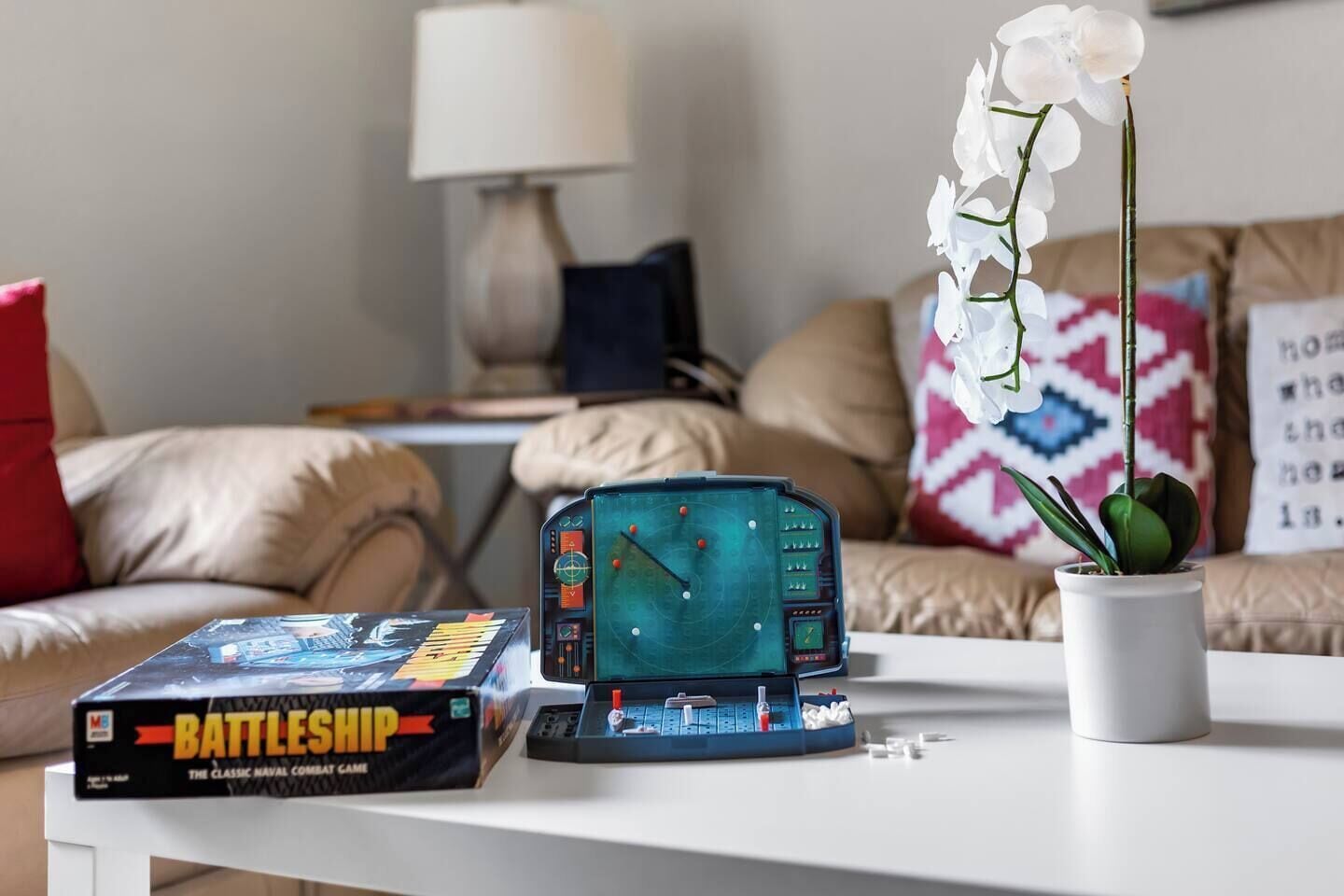 A Battleship game box, a board game, and a potted orchid on a white table in a living room.
