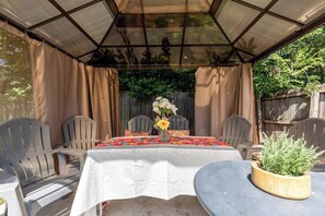 Ein überdachter Patio-Bereich mit einem für zwei Personen gedecktem Tisch, umgeben von Stühlen und einem Blumentopf auf dem Tisch.