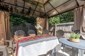 En overdækket patio med et bord dækket til to, omgivet af stole, samt et lille rundt bord med en plante.