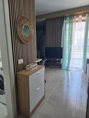 A modern living room with a wooden cabinet, a television, and a large window with curtains. - Sea & Sun Studio! A warm place for your holidays! 300 m from the Sea. (Golem)
