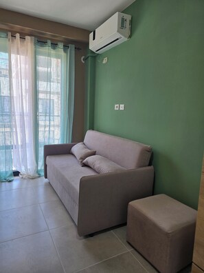 Une pièce avec un canapé beige, un pouf assorti, un mur vert et une fenêtre ornée de rideaux légers.