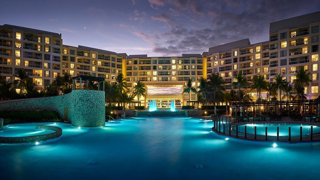 Ein Hotel mit mehreren Etagen, einem zentral gelegenen Poolbereich mit Springbrunnen und Palmen.