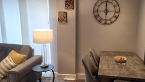 Uma sala de estar com sofá, uma mesinha de apoio com abajur, e um espaço de refeições com mesa e cadeiras.