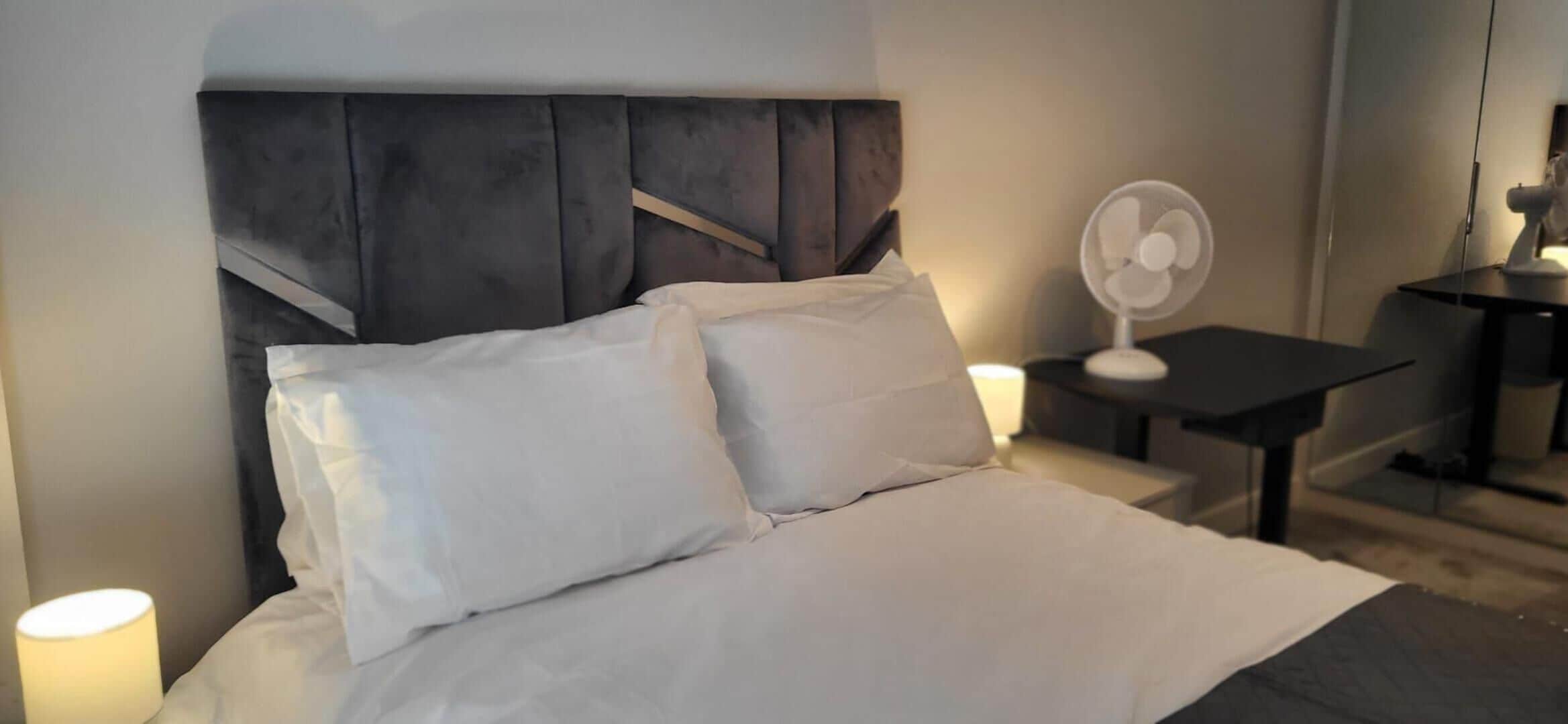 A hotel room with a bed, bedside lamps, a desk with a chair, and a standing fan.