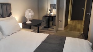 Quarto de hotel com cama, mesa de cabeceira com luminária e um ventilador.