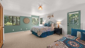 Um quarto com uma cama grande, uma cama menor, uma mesa de cabeceira, uma luminária, um objeto decorativo na parede e uma porta.