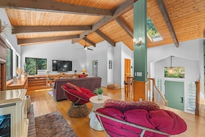 A spacious living area with a high ceiling, wooden beams, a television, a sofa, a dining table, and a staircase leading to an upper level. - 3BR Gualala Forest Retreat | Game Room, Soaking Tub & ADU Suite (Mendocino County)