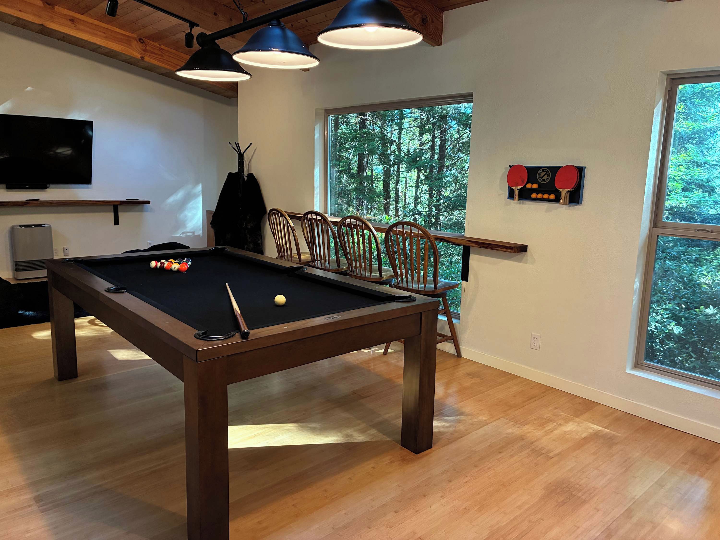 A pool table with balls, a television, and a window with a view of trees.