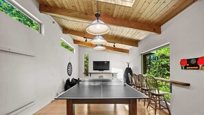 Uma mesa de ping pong em um quarto com piso de madeira, teto com vigas aparentes e uma televisão na parede.