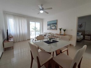 Ein modernes Wohnzimmer mit Essbereich, Deckenventilator, großem Fenster mit Vorhängen, einer Couch, einem Couchtisch und einem Fernseher.