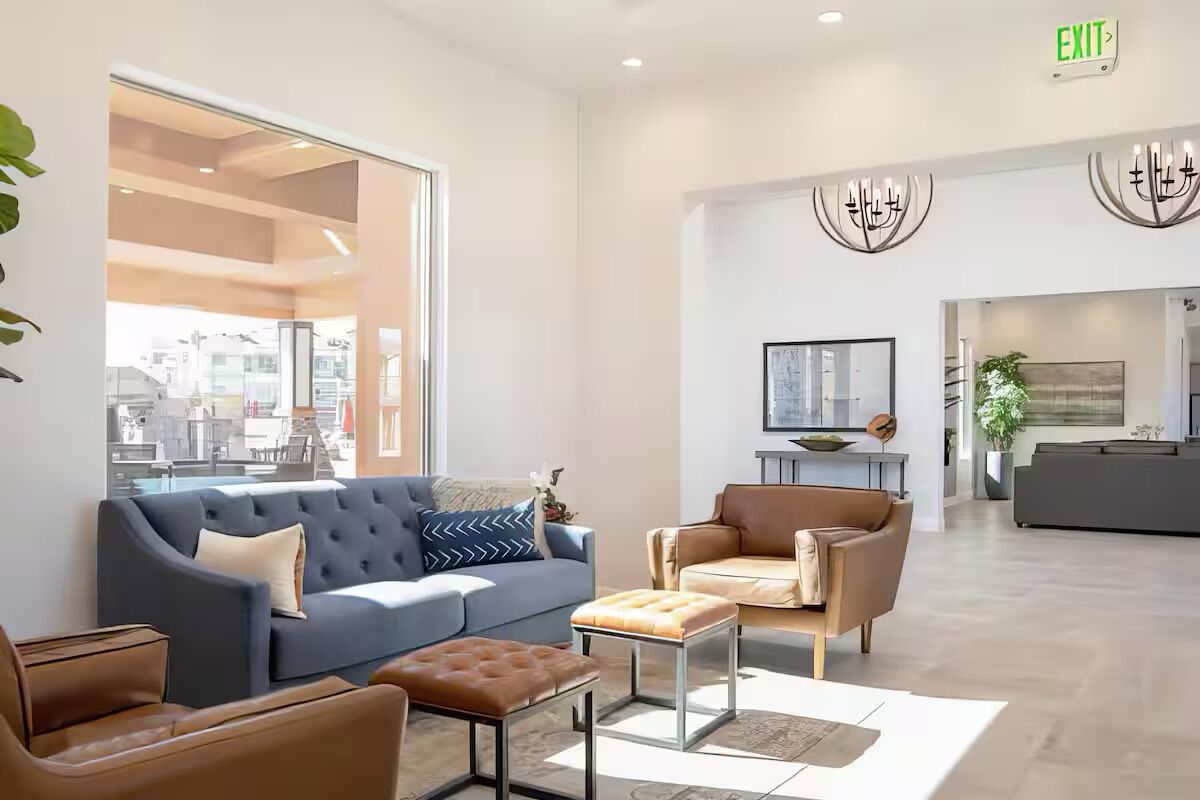 A modern hotel lobby with a blue sofa, a brown armchair, and a coffee table. There is a large window providing a view of the city, and an exit sign is visible on the wall.