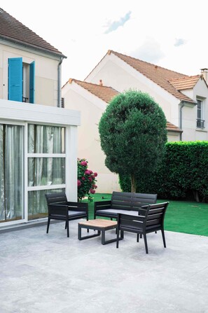 An outdoor seating area with a table and chairs, a potted tree, and a view of a residential building with a tiled roof.