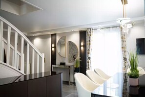 A modern interior with a staircase, a dining area with a white table and chairs, a kitchen area, and a large window with curtains.