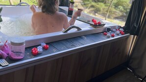 En person i ett bubbelbad med en flaska och ett glas, omgiven av träväggar och med utsikt över grönskan.