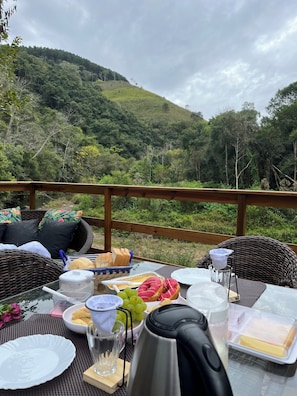 An outdoor dining area with wicker furniture, a table set with food and drinks, and a view of a forested hillside. - My eucalyptus tree cabin - a little corner of paradise. (Alfredo Wagner)