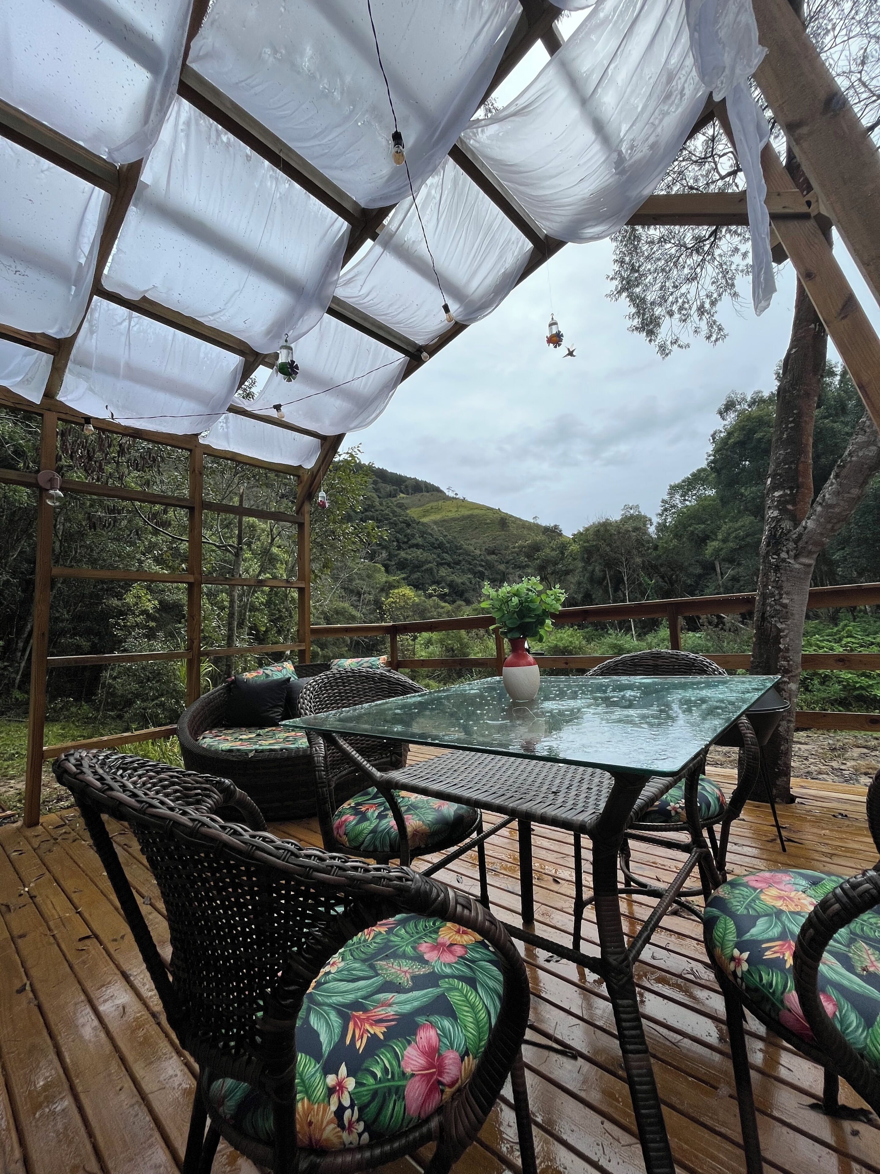 Une terrasse extérieure avec une table en verre et des chaises en osier, entourée d’une plateforme en bois et d’un auvent blanc.