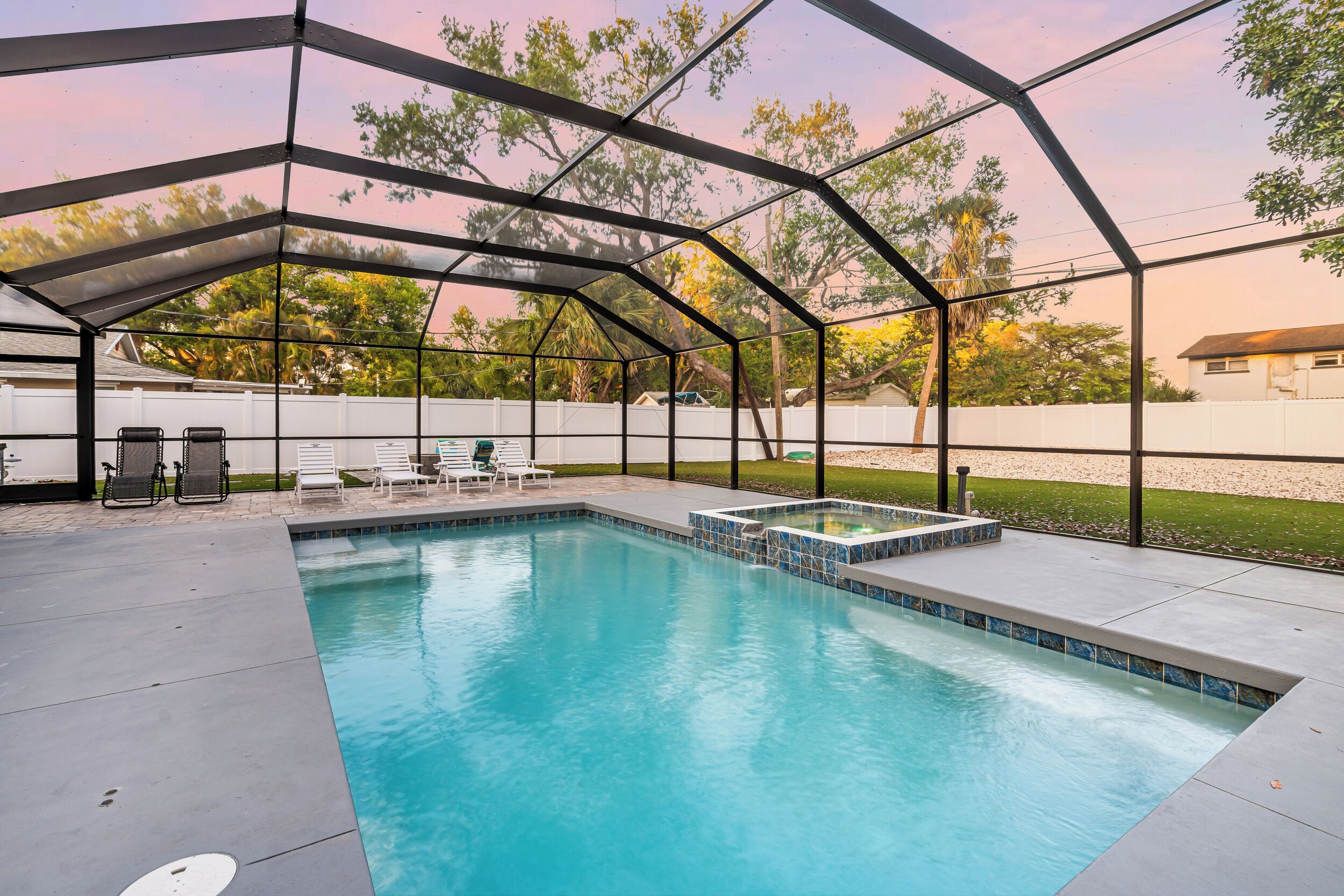 A swimming pool with a covered area, surrounded by a white fence, and trees in the background.