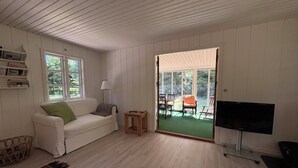 Quarto com sofá branco, uma mesinha, televisão e uma porta de correr de vidro que dá para uma área externa com mesa e cadeiras.