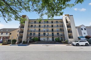 A multi-story residential building with balconies, a parking lot, and a white car parked in front. - Bayfront 1BR With Pool Near Beach (Ocean City)