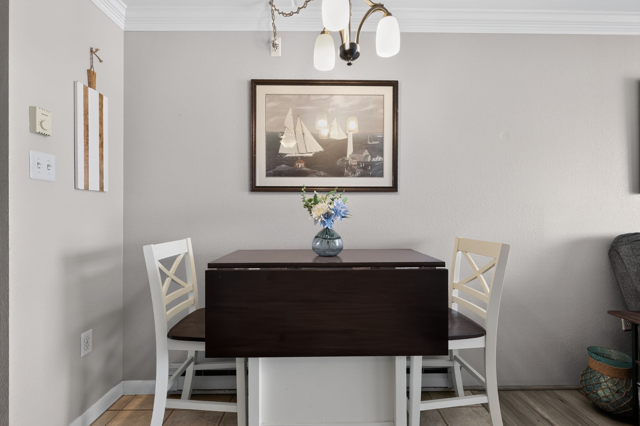 A dining area with a dark wooden table, white chairs, a vase with flowers, and a framed picture on the wall.