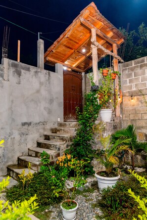 A rustic outdoor area with a wooden pergola, a concrete wall, and a set of stone steps leading to a door.