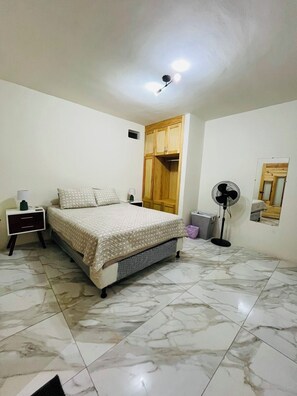 A bedroom with a bed, nightstand, wardrobe, fan, and a small window.