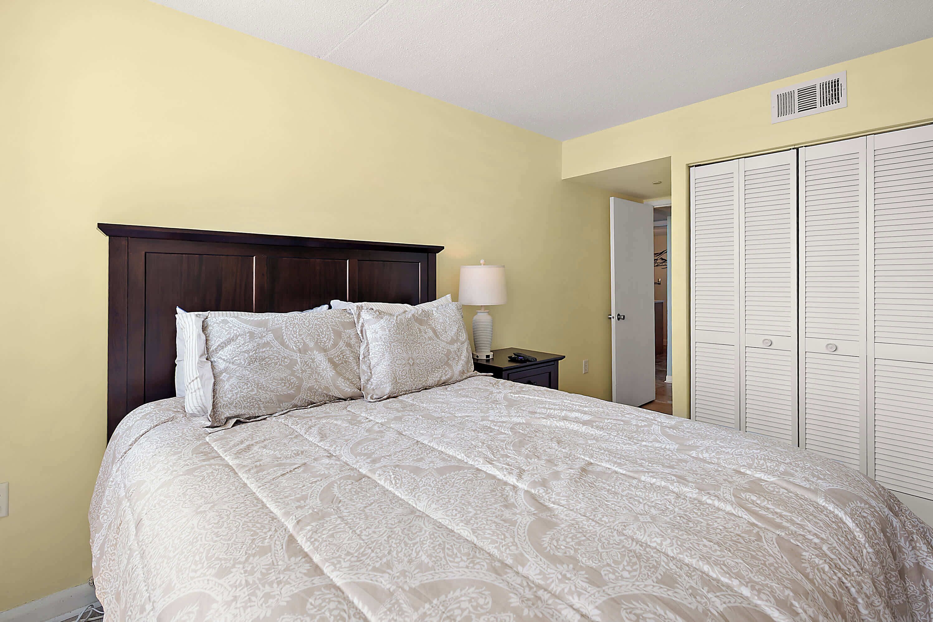 A bed with a dark headboard and multiple pillows, a bedside lamp, and a wardrobe with louvered doors.
