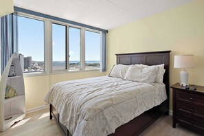 Une chambre avec une grande fenêtre, un lit avec une literie à motifs, une table de chevet avec une lampe et une commode.