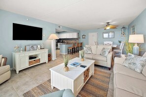 A living room with a flat-screen TV, a white coffee table, a sofa, and a dining area with chairs. - Two Bedroom Beachfront Condo with Great Amenities (Ocean City)