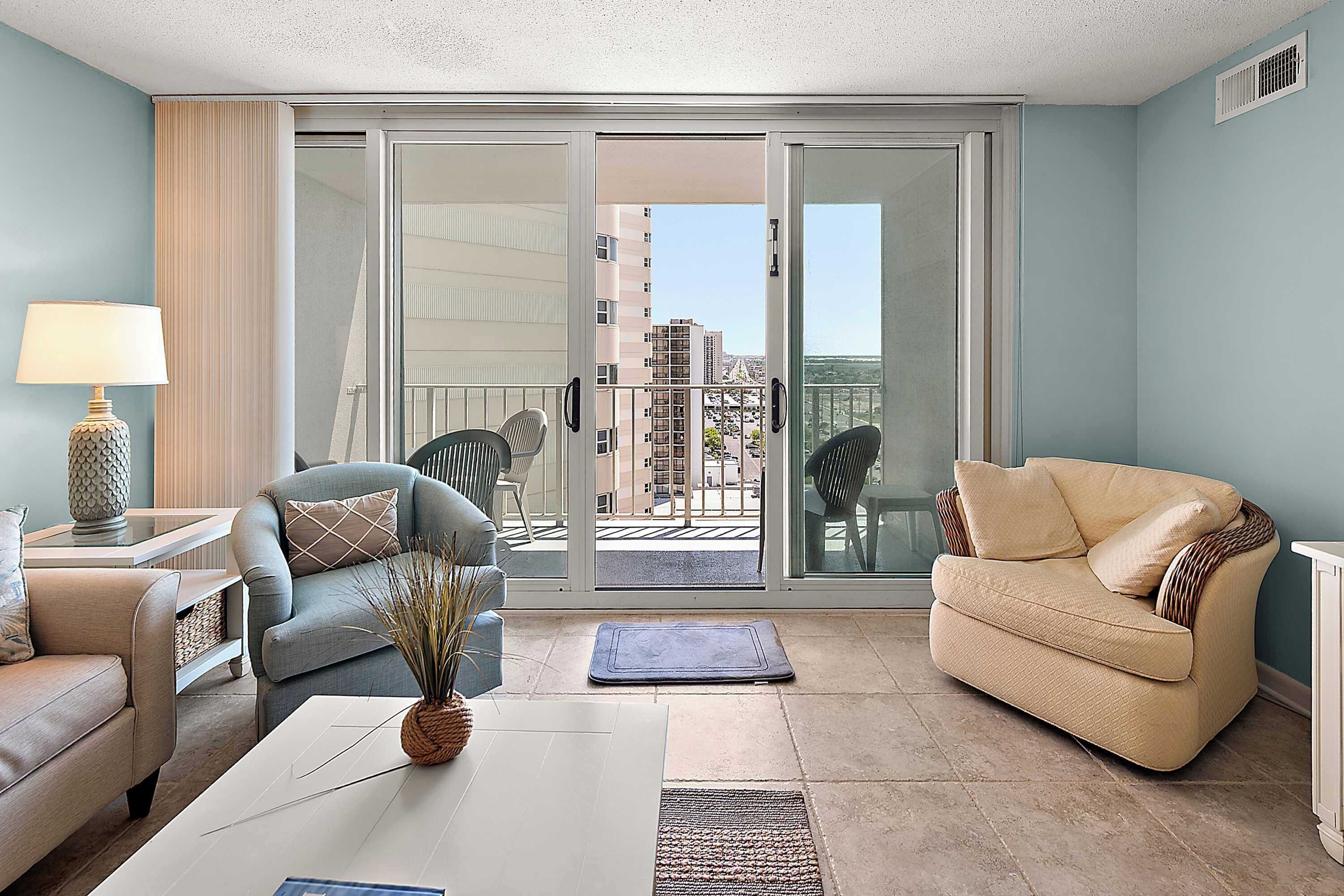 A living room with a sofa, armchair, and a coffee table. There is a view through a sliding glass door to the outside.